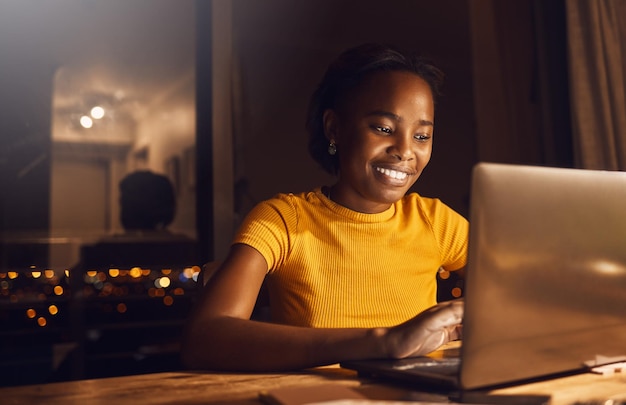 Joven mujer feliz y hermosa que trabaja y estudia por la noche para cumplir con la fecha límite del curso en línea Estudiante sonriente y positiva que usa una computadora portátil para terminar su proyecto mientras está sentada en su escritorio en casa