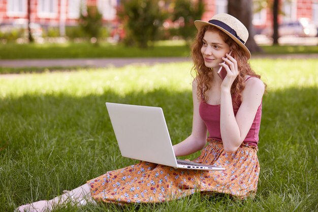 Joven mujer feliz llamando con un teléfono inteligente mientras toma un descanso entre el trabajo en la computadora portátil, sonriente estudiante hablando por teléfono celular mientras está sentado con net-book en el parque durante el tiempo de recreación