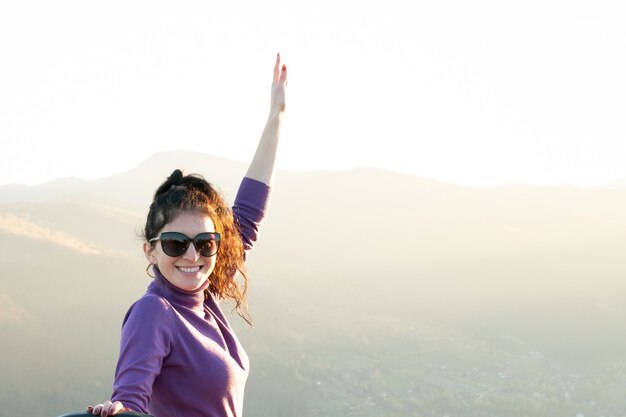 Joven mujer feliz con la mano levantada disfrutando de la cálida noche del atardecer en las montañas de verano.