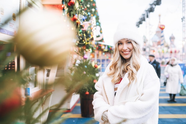 Joven mujer feliz con el pelo rizado en un sombrero de punto blanco de compras en el mercado de la feria de Navidad en la calle de invierno decorada con luces