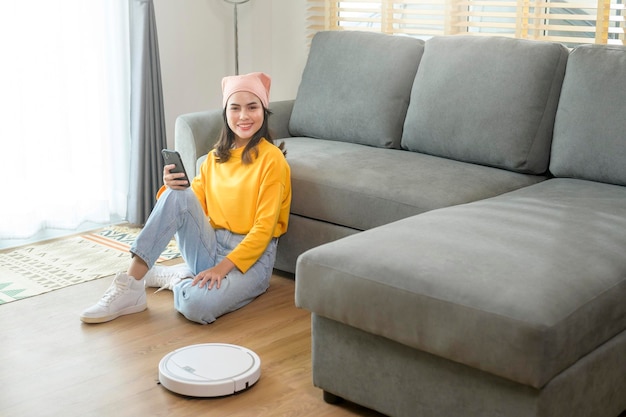Joven mujer feliz relajándose y usando un teléfono inteligente en la sala de estar mientras la aspiradora robótica funciona