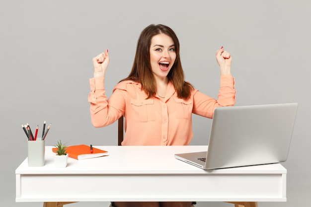 Joven mujer feliz con ropa casual pastel haciendo gesto de ganador, siéntese a trabajar en un escritorio blanco con una computadora portátil aislada en un fondo gris. Concepto de estilo de vida de carrera empresarial de logro. Simulacros de espacio de copia.