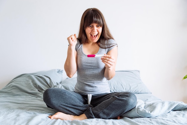 Joven mujer feliz sentada en la cama mirando una prueba de embarazo positiva