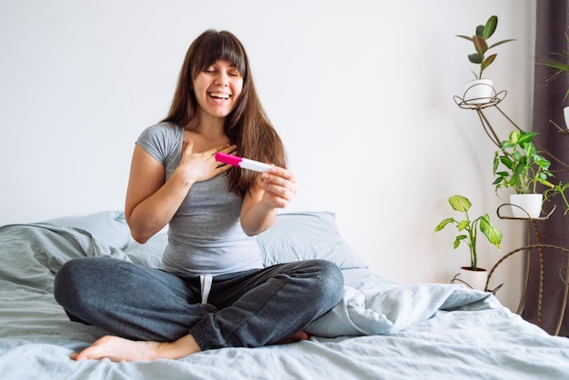 Joven mujer feliz sentada en la cama mirando una prueba de embarazo positiva