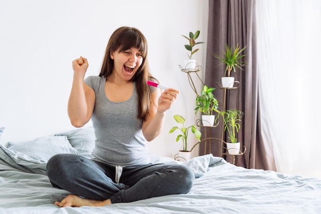 Joven mujer feliz sentada en la cama mirando una prueba de embarazo positiva