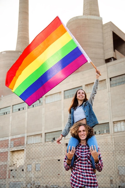 Joven mujer feliz sentada sobre los hombros de un amigo ondeando la bandera del arco iris en la demostración de los derechos LGBT Imagen vertical