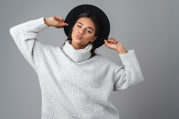 Joven mujer feliz con sombrero negro posando y haciendo beso labios aislados sobre pared gris