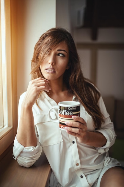 Una joven mujer feliz y sonriente está bebiendo café matutino sentada en la habitación y no puede despertarse.