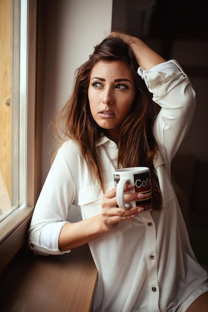 Una joven mujer feliz y sonriente está bebiendo café matutino sentada en la habitación y no puede despertarse.