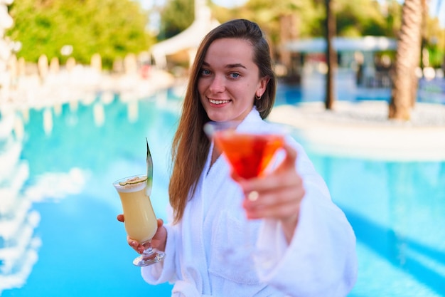 Joven mujer feliz y sonriente que disfruta de las vacaciones de verano con refrescantes cócteles de piña colada y aperol junto a la piscina en el resort todo incluido