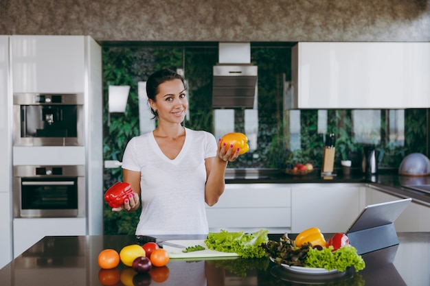 La joven mujer feliz sosteniendo verduras en las manos en la cocina con el portátil sobre la mesa