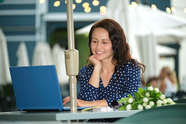 Joven mujer feliz trabajando de forma remota en una computadora portátil sentada en una mesa de restaurante al aire libre Concepto de hacer negocios en línea