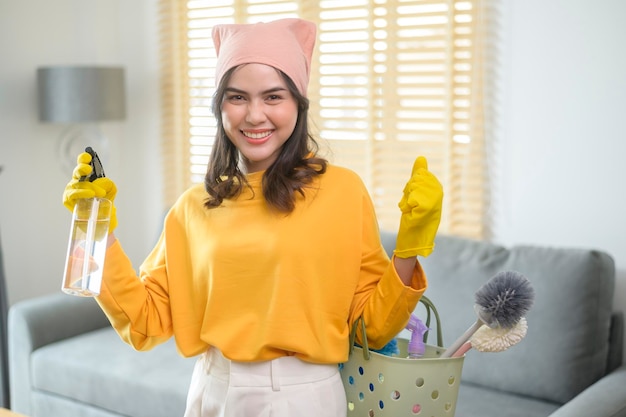 Joven mujer feliz usando guantes amarillos y sosteniendo una canasta de artículos de limpieza en la sala de estar