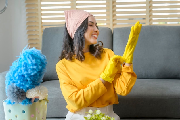 Joven mujer feliz usando guantes amarillos y sosteniendo una canasta de artículos de limpieza en la sala de estar