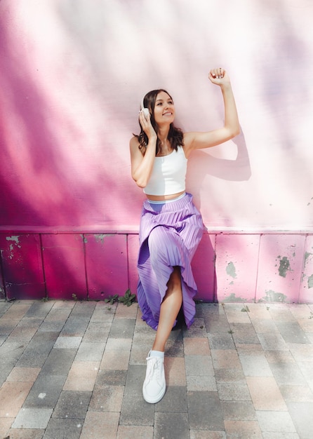 Una joven mujer feliz vestida con un top blanco y auriculares con falda morada escuchando música bailando en