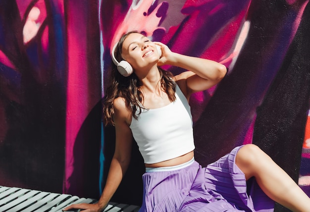 Una joven mujer feliz vestida con un top blanco y auriculares con falda morada escuchando música bailando en