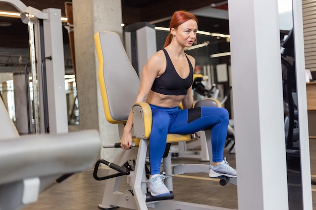 Una joven mujer fitness pelirroja entrena los músculos de las piernas en una máquina de ejercicios en un gimnasio moderno