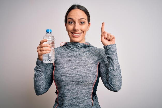 Joven mujer fitness con ropa deportiva bebiendo agua de una botella de plástico sorprendida con una idea o pregunta señalando con el dedo con la cara feliz número uno