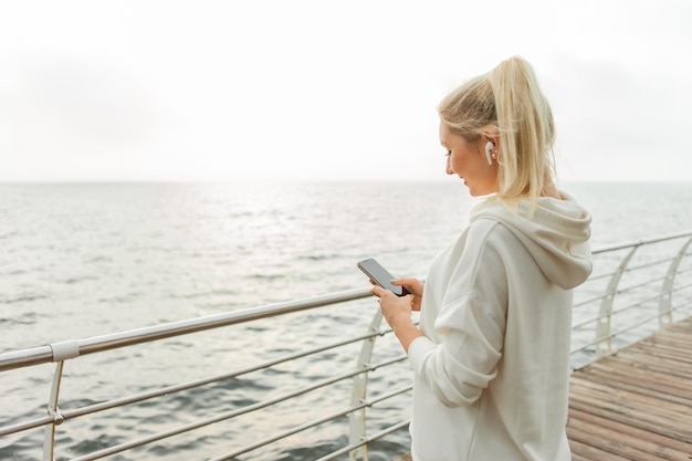 Joven mujer fitness rubia con una capucha blanca usa un teléfono inteligente y escucha música con auriculares en la playa