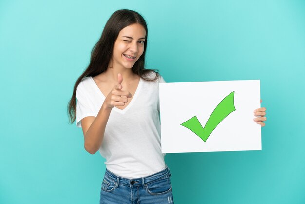 Foto joven mujer francesa aislada sobre fondo azul sosteniendo un cartel con el icono de marca de verificación verde de texto y apuntando hacia el frente