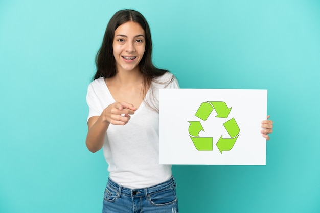 Joven mujer francesa aislada sobre fondo azul sosteniendo un cartel con el icono de reciclaje y apuntando hacia el frente