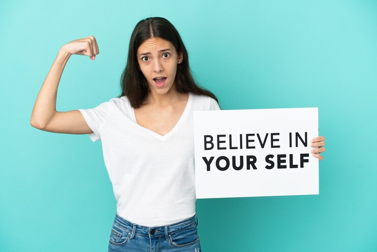 Foto joven mujer francesa aislada sobre fondo azul sosteniendo un cartel con el texto cree en ti mismo y haciendo un gesto fuerte