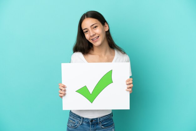 Joven mujer francesa aislada sobre fondo azul sosteniendo un cartel con texto Icono de marca de verificación verde con expresión feliz