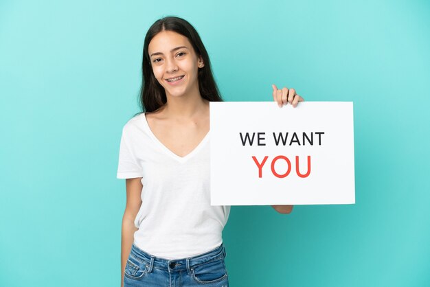 Joven mujer francesa aislada sobre fondo azul sosteniendo We Want You board con expresión feliz