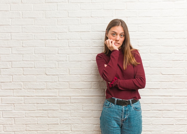 Joven mujer fresca sobre una pared de ladrillos mordiéndose las uñas, nerviosa y muy ansiosa