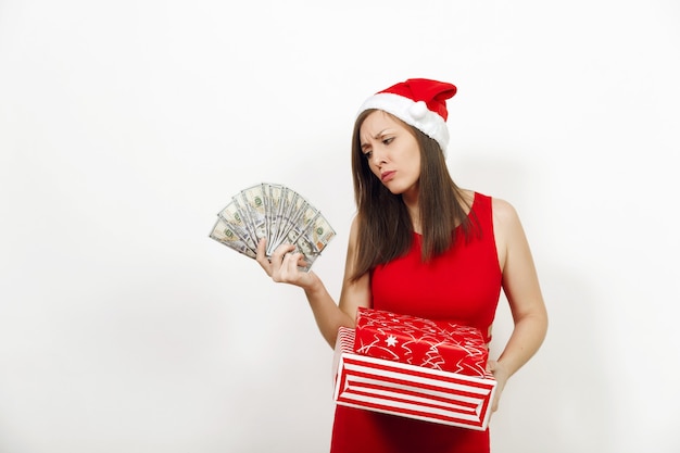 Joven mujer frustrada desconcertada en vestido rojo y sombrero de Navidad con cajas de regalo y billetes de dinero sobre fondo blanco. Niña de Santa con presente y efectivo aislado. Concepto de vacaciones de año nuevo 2018.