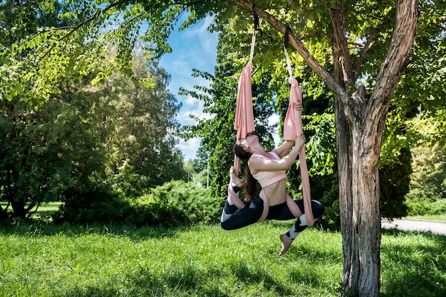 Joven mujer fuerte con una cintura delgada con brazos fuertes que se dedica a ejercicios en una hamaca de aire en la naturaleza Concepto de yoga con mosca al aire libre