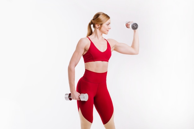Joven mujer fuerte y deportiva en tela roja haciendo ejercicios con pesas sobre fondo blanco.