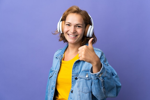 Joven mujer georgiana aislada en la pared púrpura escuchando música y con el pulgar hacia arriba