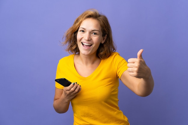 Joven mujer georgiana aislada sobre fondo púrpura mediante teléfono móvil mientras hace Thumbs up