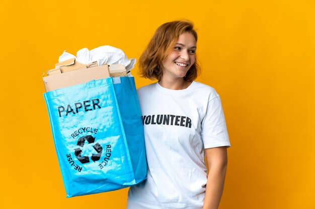 Joven mujer georgiana sosteniendo una bolsa de reciclaje llena de papel para reciclar mirando hacia el lado y sonriendo