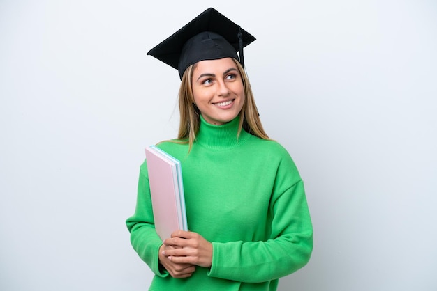 Joven mujer graduada universitaria aislada sobre fondo blanco mirando hacia el lado