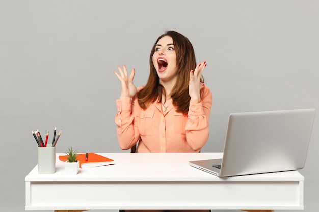 Joven mujer gritando conmocionada con ropa pastel mirando a un lado extendiendo el trabajo manual en el escritorio con una computadora portátil aislada en un fondo gris. Concepto de estilo de vida de carrera empresarial de logro. Simulacros de espacio de copia.