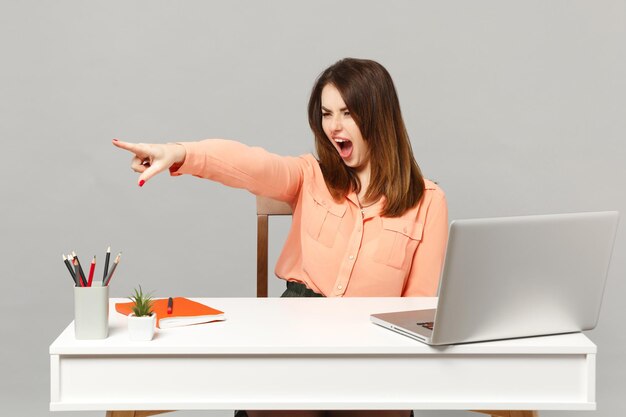 Joven mujer gritando con ropa pastel jurando señalar con el dedo índice a un lado siéntate a trabajar en un escritorio blanco con una computadora portátil pc aislada en un fondo gris. Logro el concepto de carrera empresarial. Simulacros de espacio de copia.