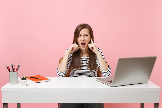 Joven mujer gruñona irritada no quiere escuchar cubriéndose los oídos con el dedo sentarse a trabajar en el escritorio blanco con una computadora portátil contemporánea