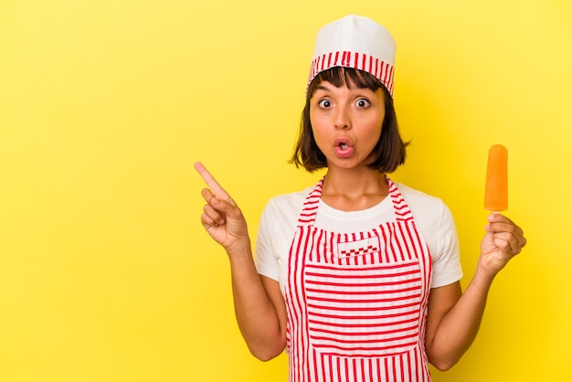 Joven mujer heladera de raza mixta sosteniendo un helado aislado sobre fondo amarillo apuntando hacia el lado