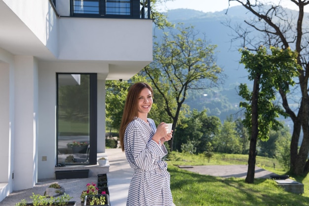 Joven mujer hermosa en una bata de baño disfrutando del café de la mañana frente a su villa de lujo