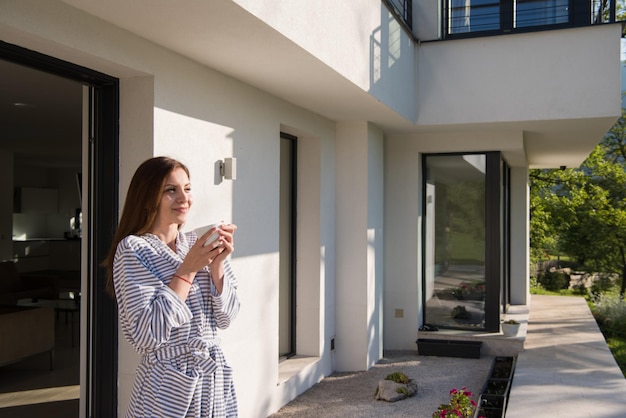 joven mujer hermosa en una bata de baño disfrutando del café de la mañana frente a su villa de lujo