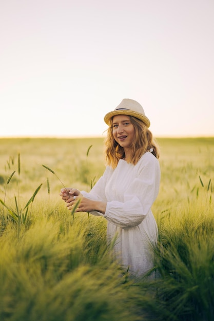 joven mujer hermosa con cabello largo rubio en un vestido blanco en un sombrero de paja recoge flores en un campo de trigo. Pelo volador al sol, verano. Hora de los soñadores, atardecer dorado.