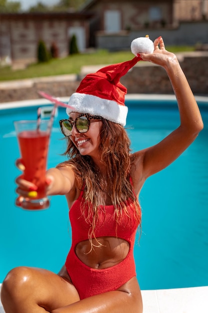 Joven mujer hermosa cerca de la piscina con sombrero de Santa Claus celebrando Año Nuevo y Navidad en un país cálido con una copa de cóctel.