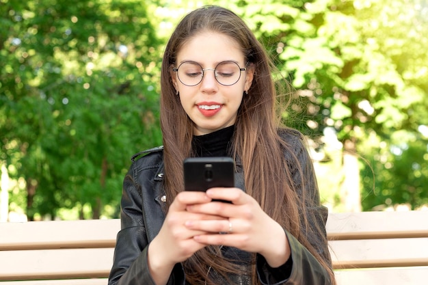 Joven mujer hermosa con una chaqueta de cuero negro escribe un mensaje o juega o mira videos o noticias de las redes sociales por teléfono sentada en un banco en un hermoso parque verde de verano o primavera