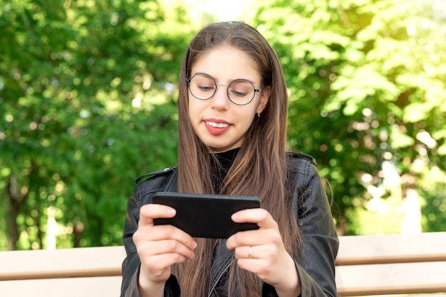 Joven mujer hermosa con una chaqueta de cuero negro escribe un mensaje o juega o mira videos o noticias de las redes sociales por teléfono sentada en un banco en un hermoso parque verde de verano o primavera