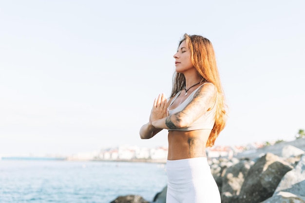 Joven mujer hermosa en forma con cabello largo en ropa deportiva blanca practica yoga y meditación en el mar