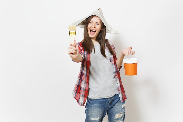 Joven mujer hermosa llena de alegría con sombrero de periódico sosteniendo un cubo de pintura y mostrando el pincel en la cámara aislada sobre fondo blanco. Instrumentos para la renovación de la habitación del apartamento. Reparar el concepto de hogar.