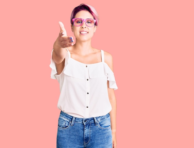 Joven mujer hermosa con el pelo rosa, vestida con ropa informal y gafas, sonriendo amablemente y ofreciendo un apretón de manos como saludo y bienvenida a un negocio exitoso