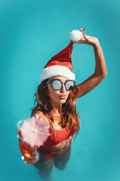 Joven mujer hermosa en la piscina con sombrero de Santa Claus, celebra Año Nuevo y Navidad en un país cálido con una copa de cóctel.
